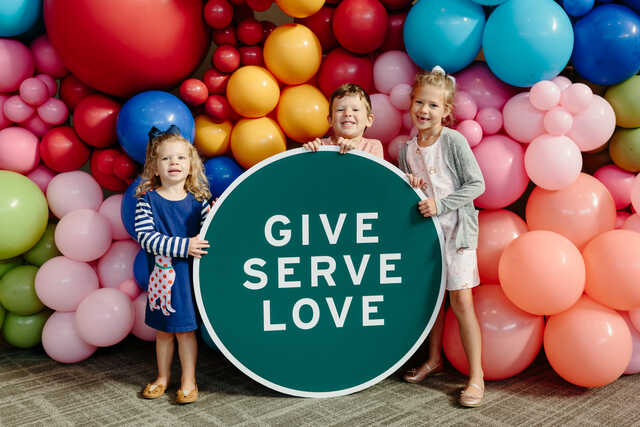 children in front of a balloon background with a give serve love sign